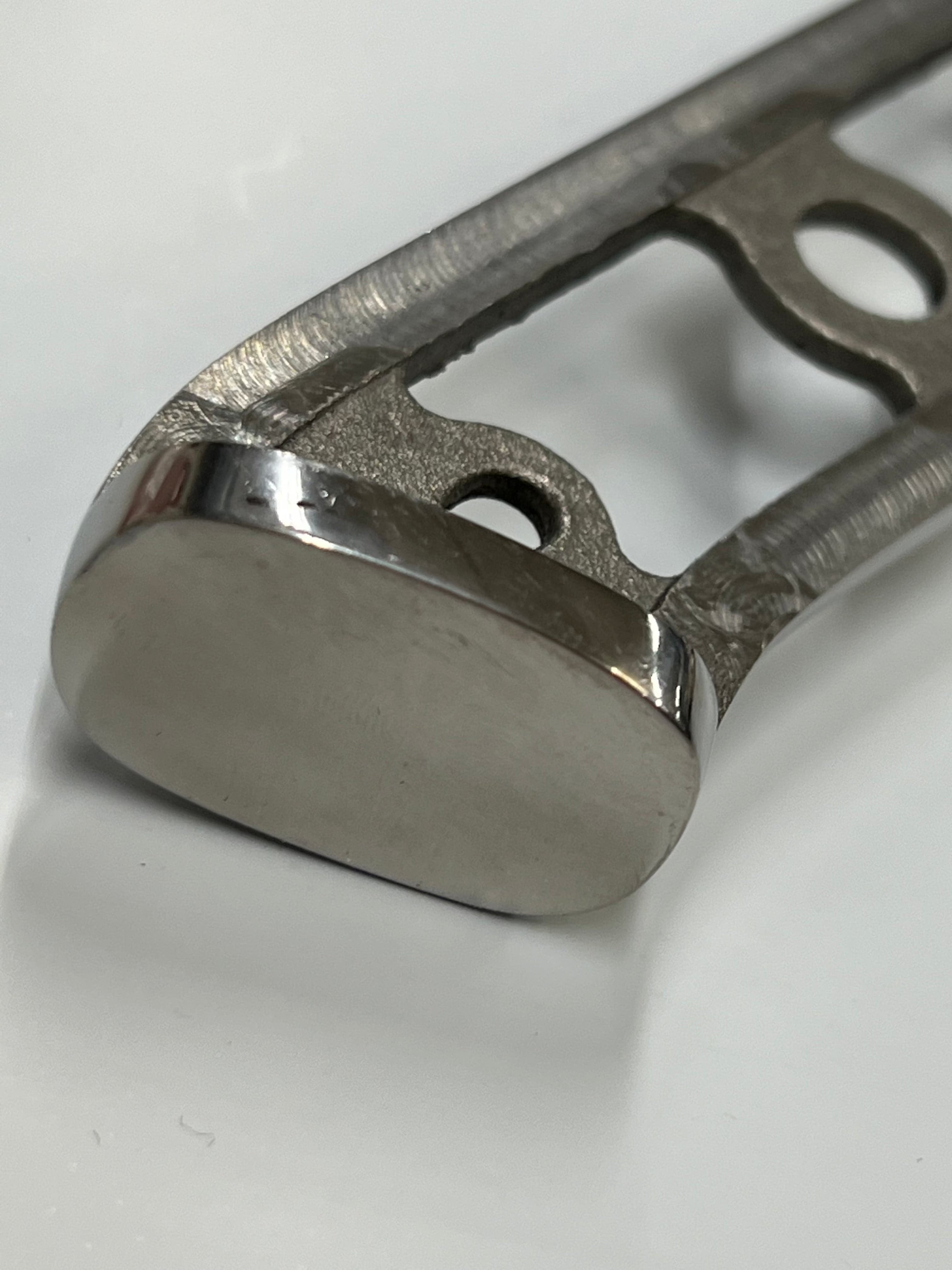 Nærbilde av en metallkomponent med en avrundet ende. Stykket har en polert, reflekterende overflate på den avrundede enden, som minner om Herring Santoku - VG10 Damask Kokkekniv 7" håndverk, og en strukturert, upolert midtseksjon med flere presise utskjæringer, inkludert sirkulære hull. Komponenten hviler på en vanlig hvit overflate.